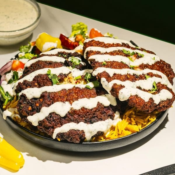 Crispy fried chicken pieces drizzled with white sauce, served over yellow rice with salad and a side of sauce.