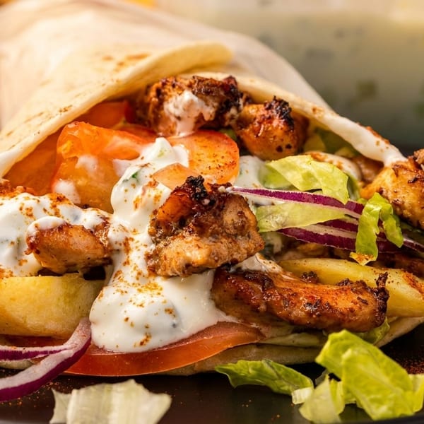A chicken shawarma wrap with fries, lettuce, tomatoes, onions, and white sauce, served with a lemon wedge.